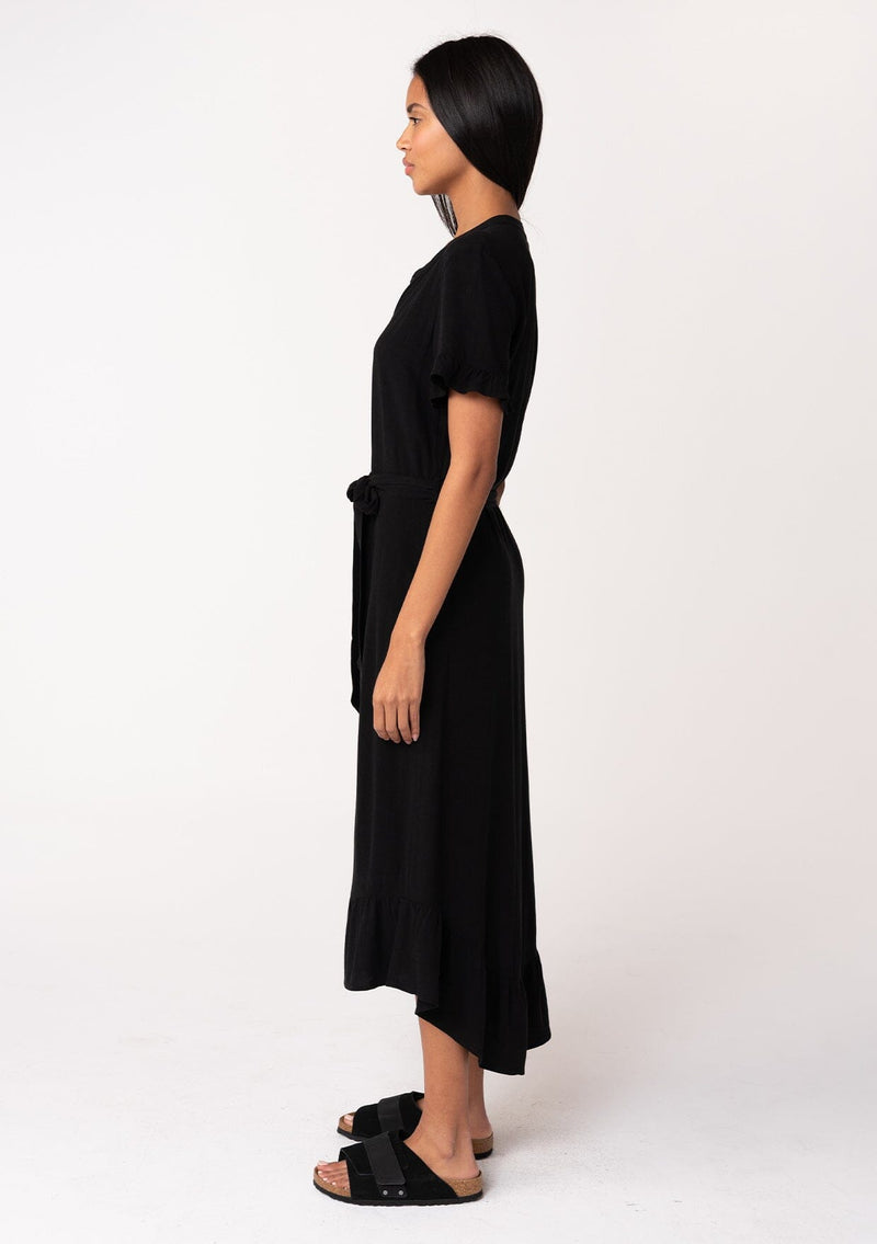 [Color: Black] A side facing image of a model wearing a black mid length shirt dress in a linen blend. Featuring short sleeves with a ruffled trim, a button up front, an adjustable waist tie, and a ruffled hemline.
