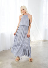 [Color: Chambray/Ivory] A model wearing a chambray blue and ivory striped maxi dress. With a halter neckline, a back tie neck with keyhole detail, an elastic waist, and a tiered skirt. 