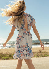 [Color: Ivory/Blue] A back facing image of a blonde model at the beach wearing a sheer floral print mini dress. With short flounce sleeves, a braided trim throughout, an open back with braided rope tie, and a smocked elastic waist.