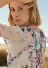 [Color: Ivory/Blue] A close up side facing image of a blonde model at the beach wearing a sheer floral print mini dress. With short flounce sleeves, a braided trim throughout, an open back with braided rope tie, and a smocked elastic waist.