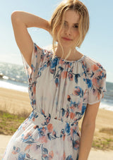 [Color: Ivory/Blue] A close up front facing image of a blonde model at the beach wearing a sheer floral print mini dress. With short flounce sleeves, a braided trim throughout, an open back with braided rope tie, and a smocked elastic waist.