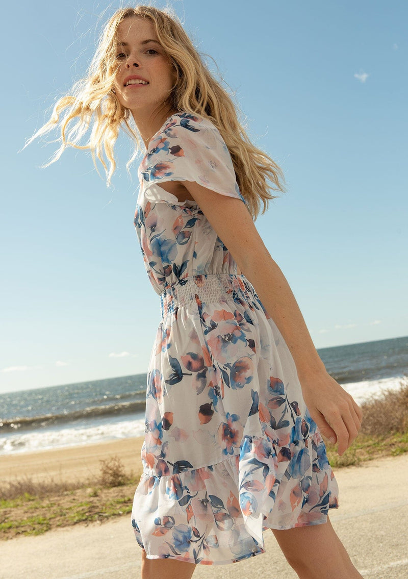 [Color: Ivory/Blue] A side facing image of a blonde model at the beach wearing a sheer floral print mini dress. With short flounce sleeves, a braided trim throughout, an open back with braided rope tie, and a smocked elastic waist.