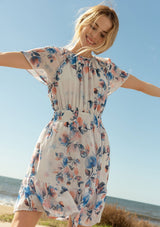 [Color: Ivory/Blue] A front facing image of a blonde model at the beach wearing a sheer floral print mini dress. With short flounce sleeves, a braided trim throughout, an open back with braided rope tie, and a smocked elastic waist.