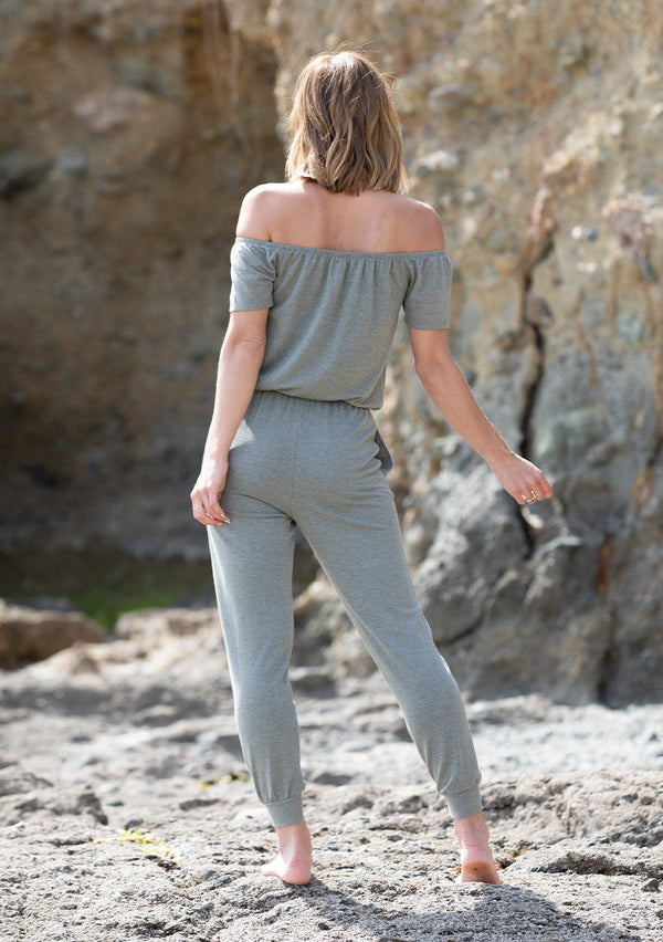 [Color: Olive] A woman standing on the beach wearing a chic off shoulder jumpsuit. Featuring a a short sleeve, an adjustable drawstring waistband, side pockets, and a slim tapered leg.
