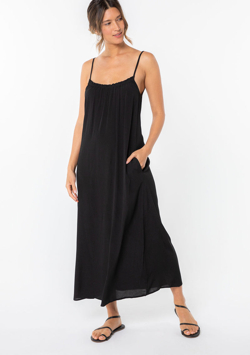 [Color: Black] A model wearing a sleeveless maxi dress. With adjustable spaghetti straps, a ruffle trimmed scooped neckline, and a v back with cutout detail.