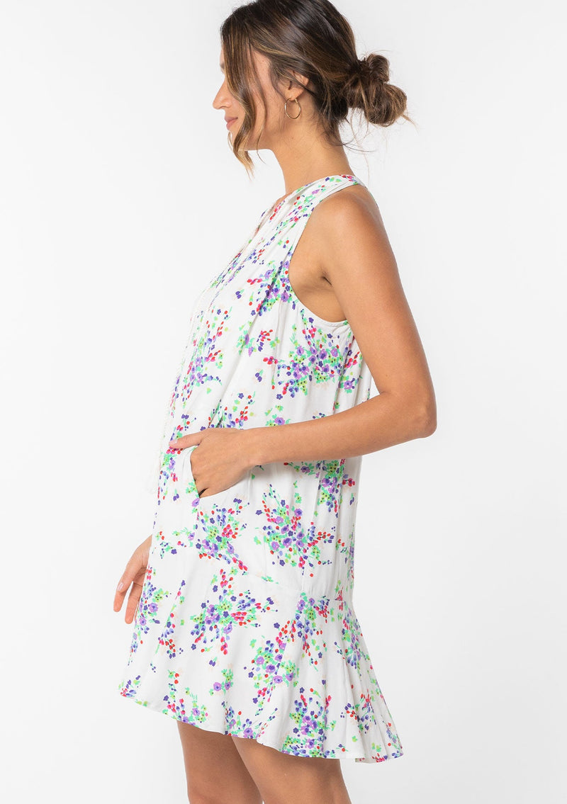 [Color: Ivory/Violet] A model wearing a white sleeveless mini dress with a purple floral print throughout. Featuring a loose and flowy tent silhouette and pleated front. 