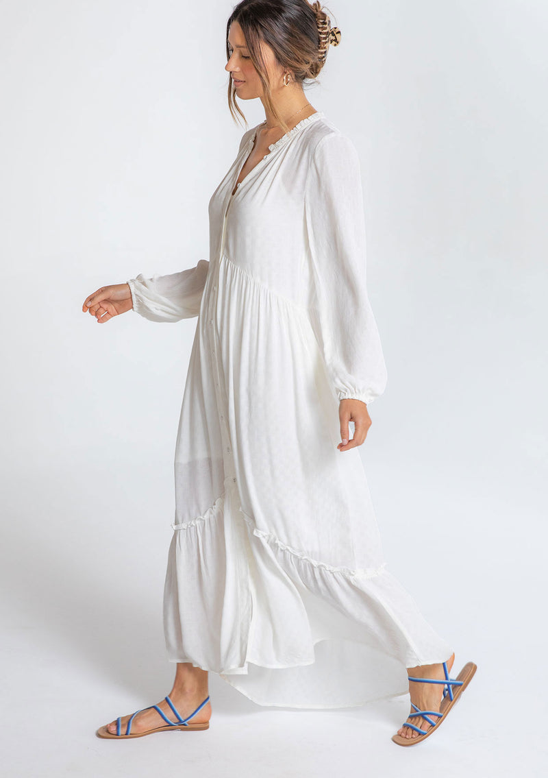 [Color: Chalk] A model wearing a timeless white maxi peasant dress with a button front, ruffled neckline, long voluminous sleeves, and tiered skirt.