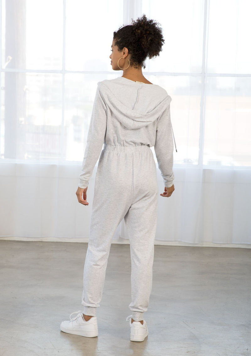 [Color: Heather Grey] A model wearing a heather grey French terry lounge jumpsuit. With long sleeves, an elastic waist, side pockets, a drawstring hoodie, and a front zip. 