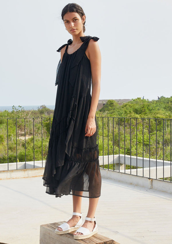 [Color: Black] A model wearing a sheer black sleeveless maxi dress. With adjustable shoulder ties, a scooped neckline, an asymmetric tiered skirt, and a wrap front.