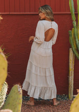 [Color: Silver] A model wearing a silver grey bohemian maxi dress in a clip dot. With an embroidered trimmed tiered skirt, a subtle high low hemline, a v neckline, a self covered button closure at the back, a sexy back cutout detail, and pin tuck details. 