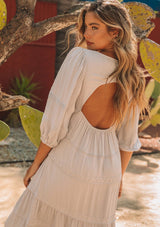 [Color: Silver] A model wearing a silver grey bohemian maxi dress in a clip dot. With an embroidered trimmed tiered skirt, a subtle high low hemline, a v neckline, a self covered button closure at the back, a sexy back cutout detail, and pin tuck details. 