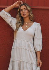 [Color: Silver] A model wearing a silver grey bohemian maxi dress in a clip dot. With an embroidered trimmed tiered skirt, a subtle high low hemline, a v neckline, a self covered button closure at the back, a sexy back cutout detail, and pin tuck details. 