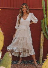 [Color: Silver] A model wearing a silver grey bohemian maxi dress in a clip dot. With an embroidered trimmed tiered skirt, a subtle high low hemline, a v neckline, a self covered button closure at the back, a sexy back cutout detail, and pin tuck details. 