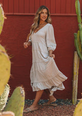 [Color: Silver] A model wearing a silver grey bohemian maxi dress in a clip dot. With an embroidered trimmed tiered skirt, a subtle high low hemline, a v neckline, a self covered button closure at the back, a sexy back cutout detail, and pin tuck details. 