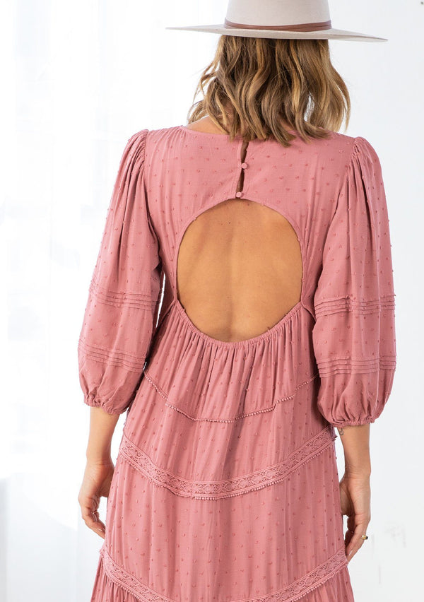 [Color: Terracotta] A model wearing a pink bohemian maxi dress in a clip dot. With an embroidered trimmed tiered skirt, a subtle high low hemline, a v neckline, a self covered button closure at the back, a sexy back cutout detail, and pin tuck details. 