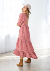 [Color: Terracotta] A model wearing a pink bohemian maxi dress in a clip dot. With an embroidered trimmed tiered skirt, a subtle high low hemline, a v neckline, a self covered button closure at the back, a sexy back cutout detail, and pin tuck details. 