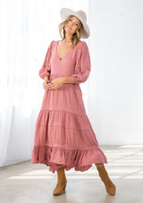 [Color: Terracotta] A model wearing a pink bohemian maxi dress in a clip dot. With an embroidered trimmed tiered skirt, a subtle high low hemline, a v neckline, a self covered button closure at the back, a sexy back cutout detail, and pin tuck details. 