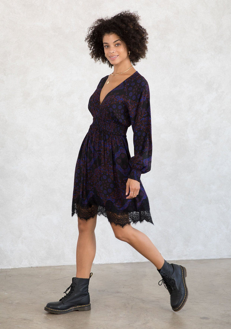 [Color: Black/Cobalt] A model wearing a black and blue paisley print bohemian mini dress. With a lace trimmed hemline, a smocked elastic waist, long voluminous sleeves with a smocked wrist cuff, and a v neckline. 