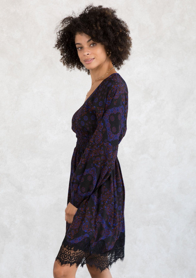 [Color: Black/Cobalt] A model wearing a black and blue paisley print bohemian mini dress. With a lace trimmed hemline, a smocked elastic waist, long voluminous sleeves with a smocked wrist cuff, and a v neckline. 