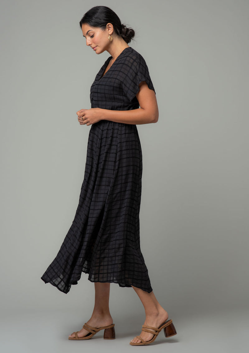 [Color: Black] A side facing image of a brunette model wearing a black cotton button front maxi dress with short sleeves and an allover textured gingham.