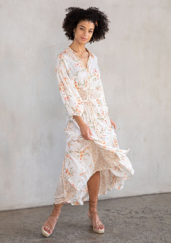[Color: Natural/Peach] A model wearing a flowy bohemian button front maxi dress in an off white and peach floral print. With three quarter length voluminous sleeves, a collared neckline, a smocked elastic waist, and a contrast floral print paneled skirt.