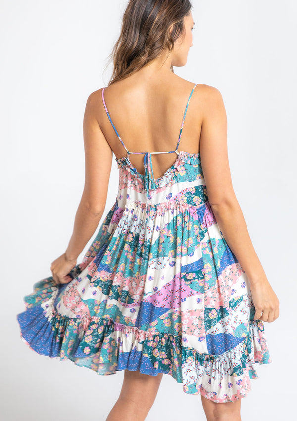 [Color: Ivory/Jade] A model wearing a white, pink, and blue patchwork floral print sleeveless mini dress with a strappy back and ruffled details. 