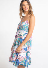 [Color: Ivory/Jade] A model wearing a white, pink, and blue patchwork floral print sleeveless mini dress with a strappy back and ruffled details. 