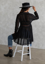 [Color: Black] A model wearing a sheer black mini dress. With lace trimmed long sleeves, a button front, a collared neckline, and a lace trimmed waist. Removable slip included. 