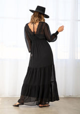 [Color: Black] A model wearing a sheer black maxi dress. With a lace trim v neckline in front and back, an open back with tassel tie closure, long sheer sleeves, a tiered skirt, and half smocked elastic waist. 