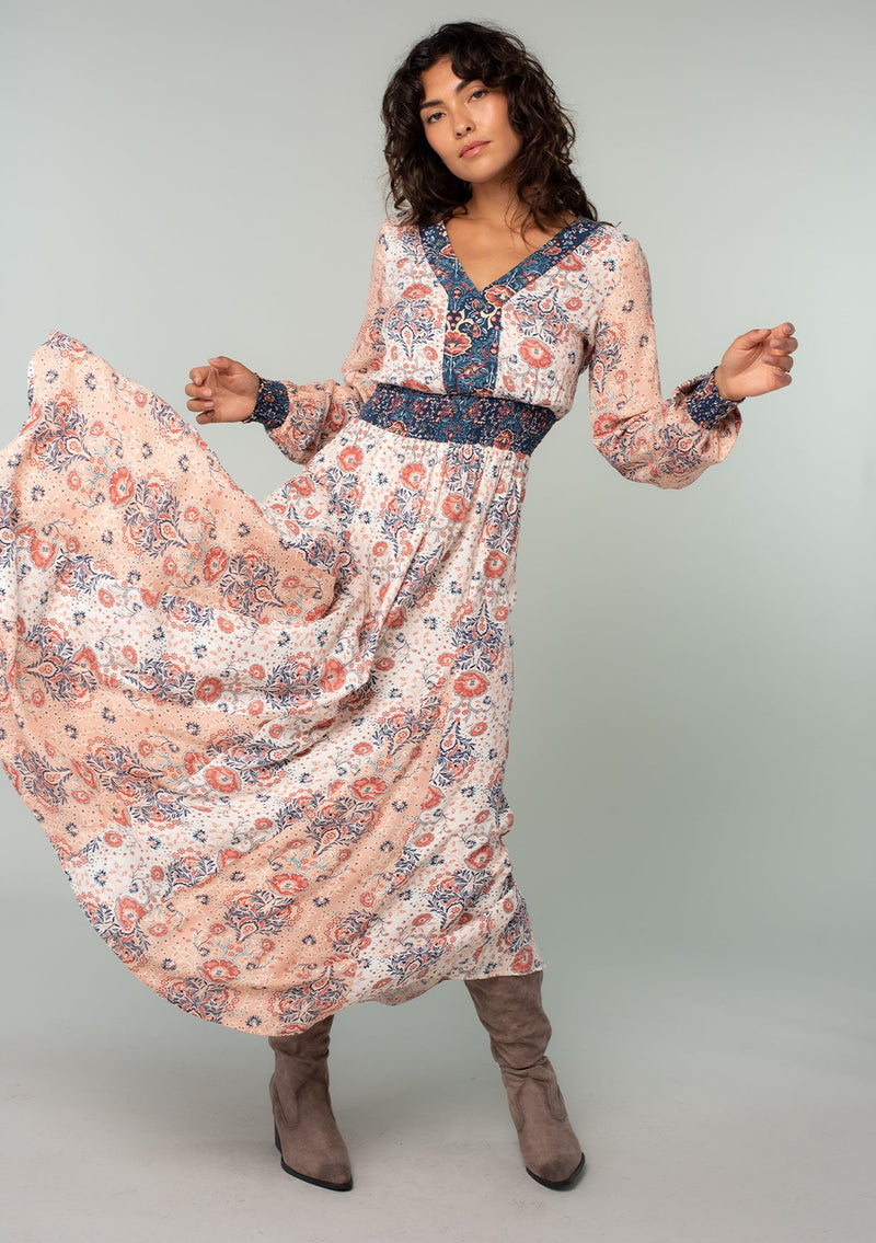 [Color: Ivory/Coral] A full body front facing image of a brunette model wearing an ivory white, coral pink, and blue mixed floral print bohemian maxi dress. With long sleeves, a v neckline, a smocked elastic waist, and a paneled flowy skirt. 