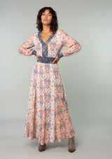 [Color: Ivory/Coral] A full body front facing image of a brunette model wearing an ivory white, coral pink, and blue mixed floral print bohemian maxi dress. With long sleeves, a v neckline, a smocked elastic waist, and a paneled flowy skirt. 