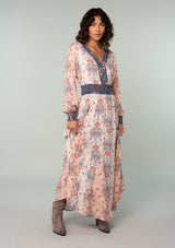 [Color: Ivory/Coral] A front facing image of a brunette model wearing an ivory white, coral pink, and blue mixed floral print bohemian maxi dress. With long sleeves, a v neckline, a smocked elastic waist, and a paneled flowy skirt. 
