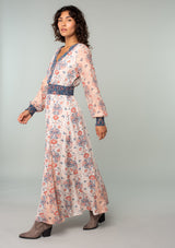 [Color: Ivory/Coral] A side facing image of a brunette model wearing an ivory white, coral pink, and blue mixed floral print bohemian maxi dress. With long sleeves, a v neckline, a smocked elastic waist, and a paneled flowy skirt. 