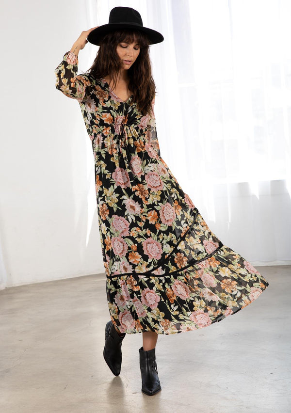 [Color: Black/Rose] A model wearing a sheer bohemian maxi dress in a large floral print. With an adjustable draw cord empire waist, long voluminous long sleeves, and lattice trim throughout. 