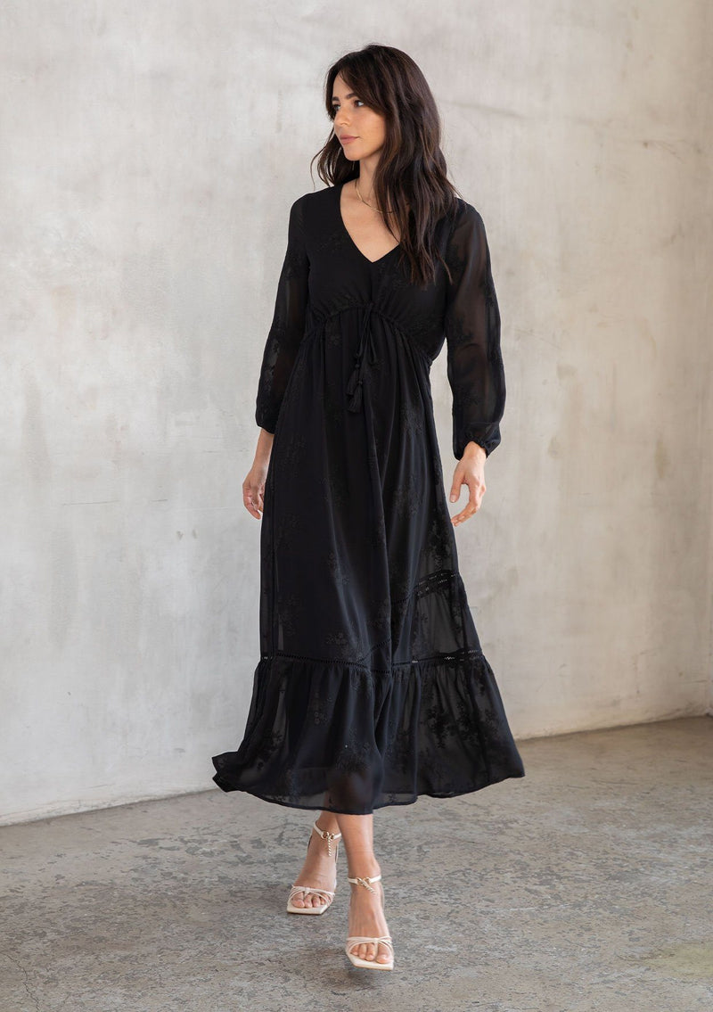 [Color: Black] A model wearing a gorgeous embroidered chiffon black maxi dress with an adjustable empire waist, tiered billowy skirt, long flattering sleeves, and lattice trim detail.