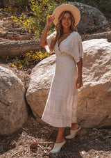 [Color: Bone] A model wearing a cream bohemian embroidered eyelet maxi dress with half length kimono sleeves, a surplice v neckline in front and back, and open back with tie detail, and a long flowy skirt.