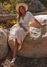 [Color: Bone] A model wearing a cream bohemian embroidered eyelet maxi dress with half length kimono sleeves, a surplice v neckline in front and back, and open back with tie detail, and a long flowy skirt.