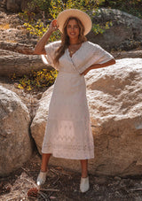 [Color: Bone] A model wearing a cream bohemian embroidered eyelet maxi dress with half length kimono sleeves, a surplice v neckline in front and back, and open back with tie detail, and a long flowy skirt.