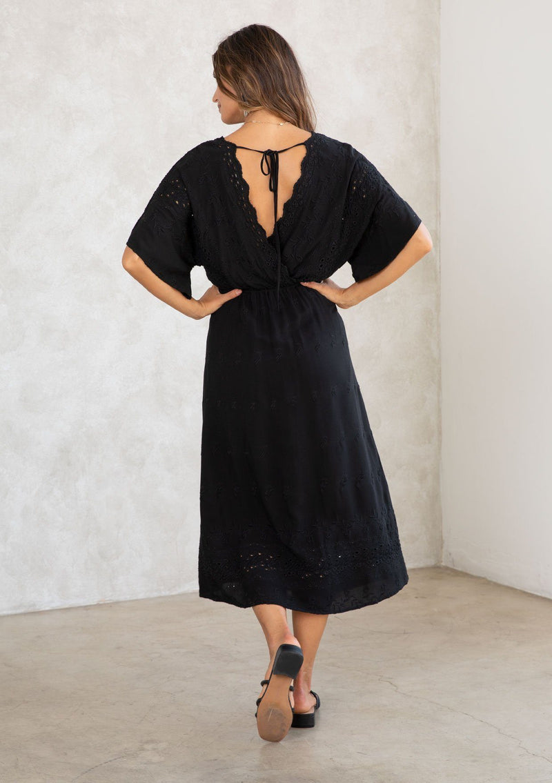 [Color: Black] A model wearing a black bohemian embroidered eyelet maxi dress with half length kimono sleeves, a surplice v neckline in front and back, and open back with tie detail, and a long flowy skirt. 