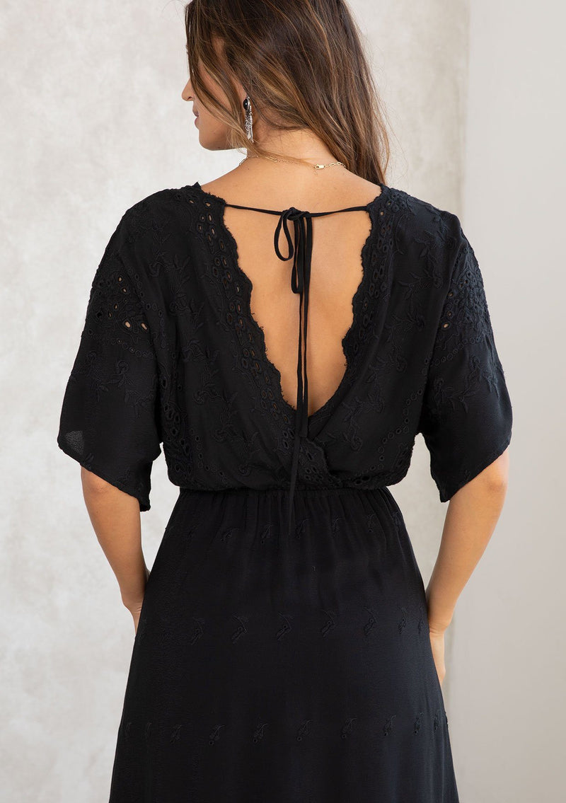 [Color: Black] A model wearing a black bohemian embroidered eyelet maxi dress with half length kimono sleeves, a surplice v neckline in front and back, and open back with tie detail, and a long flowy skirt. 