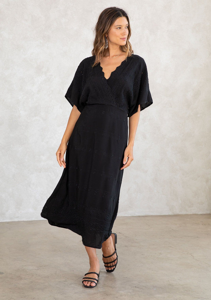 [Color: Black] A model wearing a black bohemian embroidered eyelet maxi dress with half length kimono sleeves, a surplice v neckline in front and back, and open back with tie detail, and a long flowy skirt. 