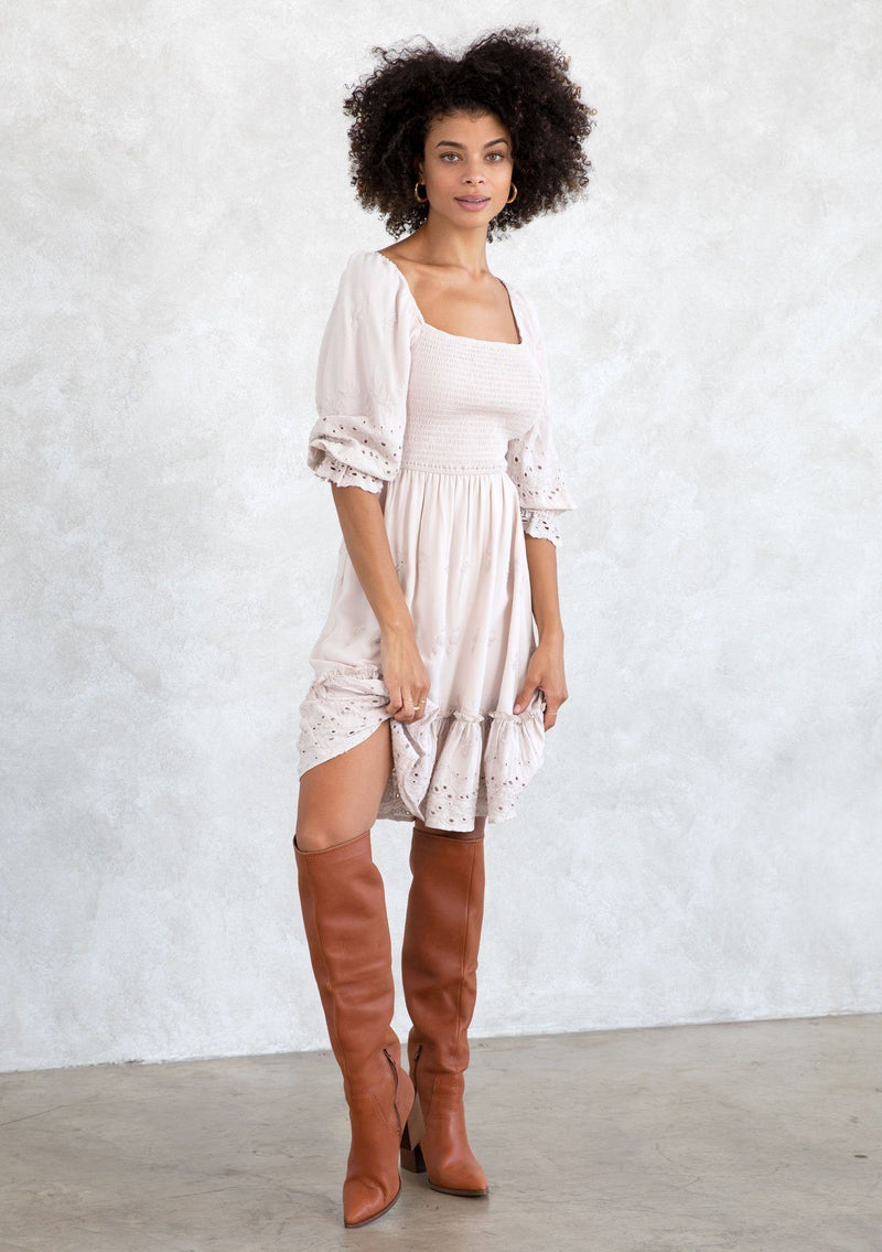 [Color: Bone] A model wearing a classic taupe bohemian mini dress with embroidered eyelet detail, a smocked bodice, half length puff sleeves, and a flowy lined skirt.