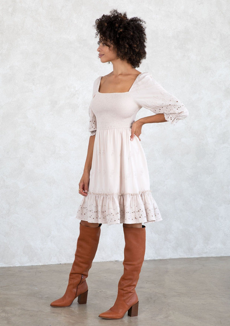 [Color: Bone] A model wearing a classic taupe bohemian mini dress with embroidered eyelet detail, a smocked bodice, half length puff sleeves, and a flowy lined skirt.