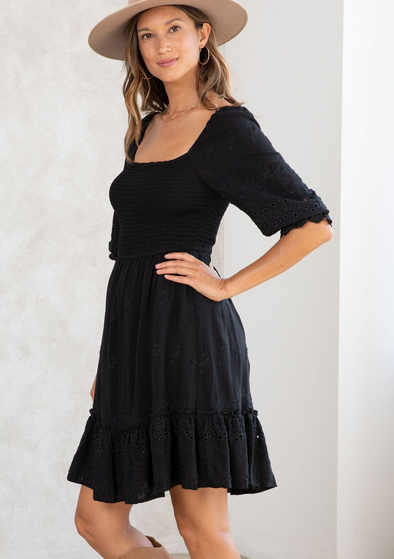 [Color: Black] A model wearing a classic black bohemian mini dress with embroidered eyelet detail, a smocked bodice, half length puff sleeves, and a flowy lined skirt. 