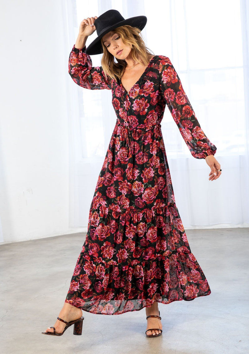 [Color: Black/Magenta] A model wearing a sheer bohemian maxi dress in a large red rose floral print. With lace trim detail throughout, a tiered skirt, an adjustable drawstring waist, and voluminous long sleeves with an elastic wrist cuff. 