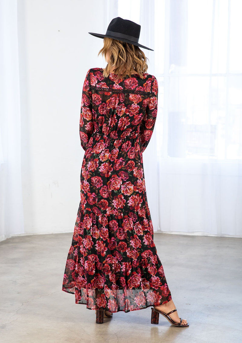 [Color: Black/Magenta] A model wearing a sheer bohemian maxi dress in a large red rose floral print. With lace trim detail throughout, a tiered skirt, an adjustable drawstring waist, and voluminous long sleeves with an elastic wrist cuff. 