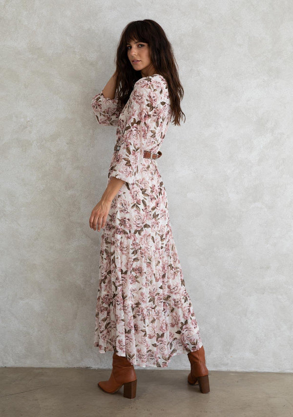 [Color: Pearl/Rose] A model wearing a sheer bohemian maxi dress in a large pink rose floral print. With lace trim detail throughout, a tiered skirt, an adjustable drawstring waist, and voluminous long sleeves with an elastic wrist cuff. 