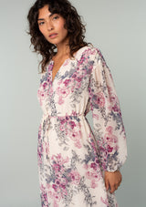 [Color: Natural/Wine] A half body side facing image of a brunette model wearing a sheer chiffon bohemian mid length dress in a natural and wine pink floral print. With voluminous long sleeves, a split v neckline, and an adjustable drawstring waist. 