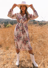 [Color: Natural/Wine] A front facing image of a blonde model wearing a sheer chiffon bohemian mid length dress in a natural and wine pink floral print. With voluminous long sleeves, a split v neckline, and an adjustable drawstring waist.