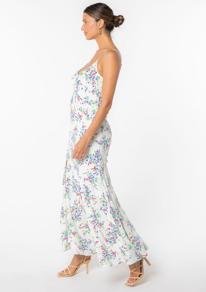 [Color: Ivory/Violet] A model wearing a white sleeveless maxi dress with purple floral print. With ruffled detail and spaghetti straps. 